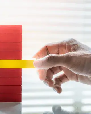 Una mà retira una peça groga d’una torre de blocs vermells, simbolitzant l’equilibri i la gestió responsable d’una organització.