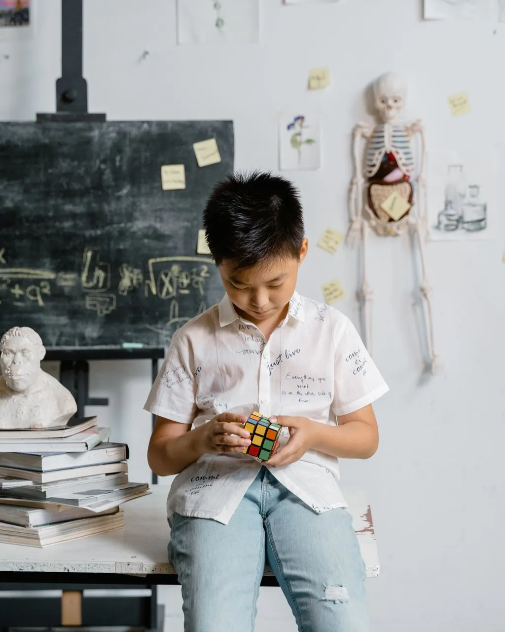 Un nen asiàtic resolent un cub de Rubik en una aula, amb llibres i materials educatius al seu voltant.
