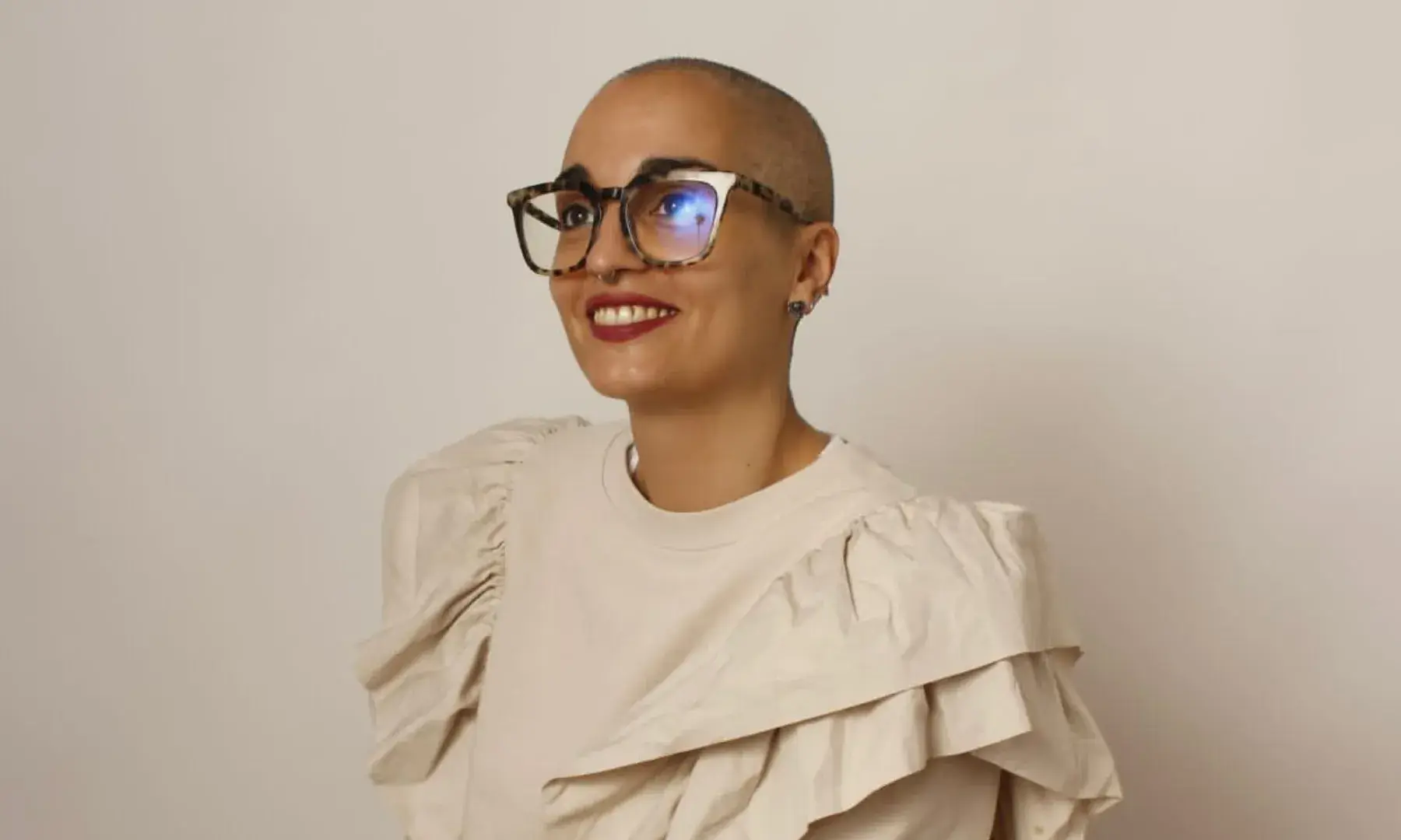 Retrat d’una dona amb alopècia i ulleres somrient sobre fons neutre.