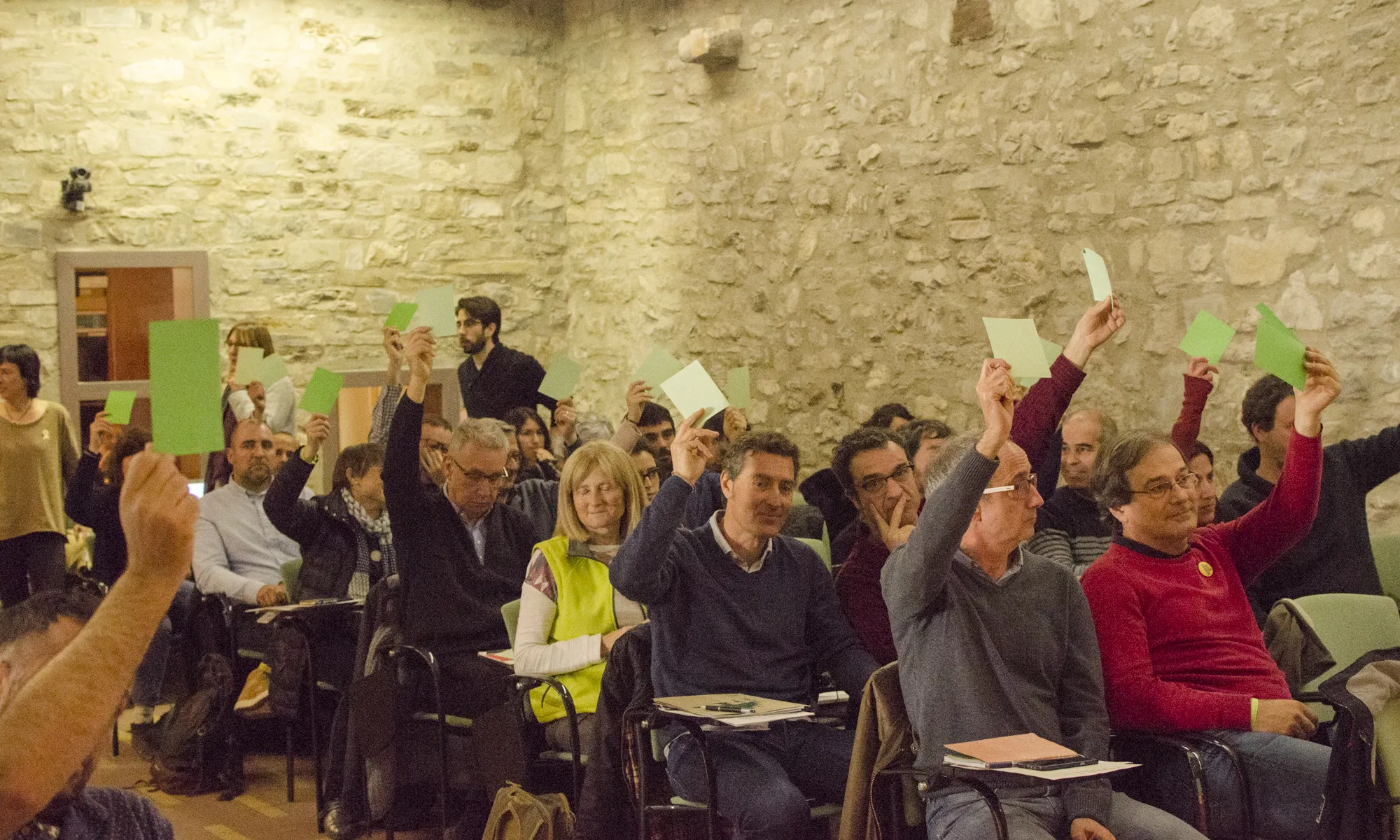 Els membres de l'assemblea estan asseguts a la sala principal del Castell de Montesquiu i voten, la majoria a favor, aixecant el braç amb cartolines verdes, sobre els diferents aspectes de la fusió de les dos entitats (XCT-XVAC).