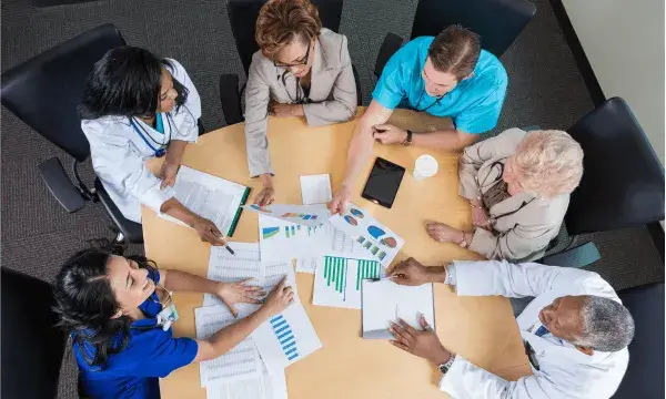 Vista zenital d’un grup de persones reunides al voltant d’una taula revisant documents i gràfics durant una reunió de treball.
