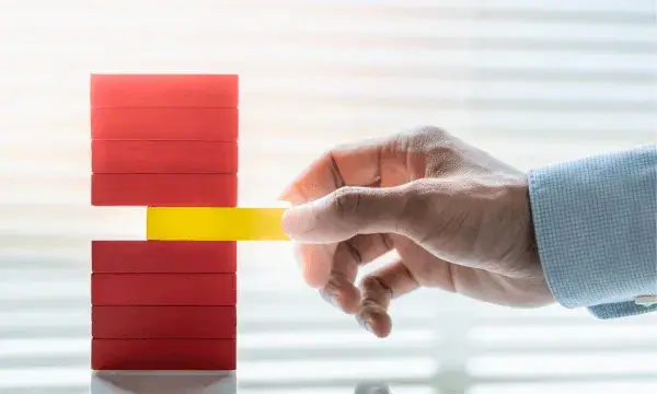 Una mà retira una peça groga d’una torre de blocs vermells, simbolitzant l’equilibri i la gestió responsable d’una organització.