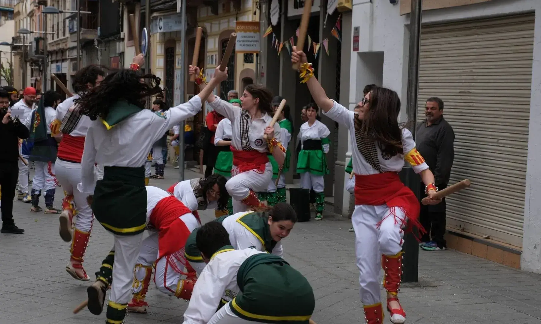 Grup de persones ballant una dansa tradicional de bastons en un carrer, vestides amb roba típica i colpejant els bastons en moviment, mentre altres participants i públic observen l’actuació.