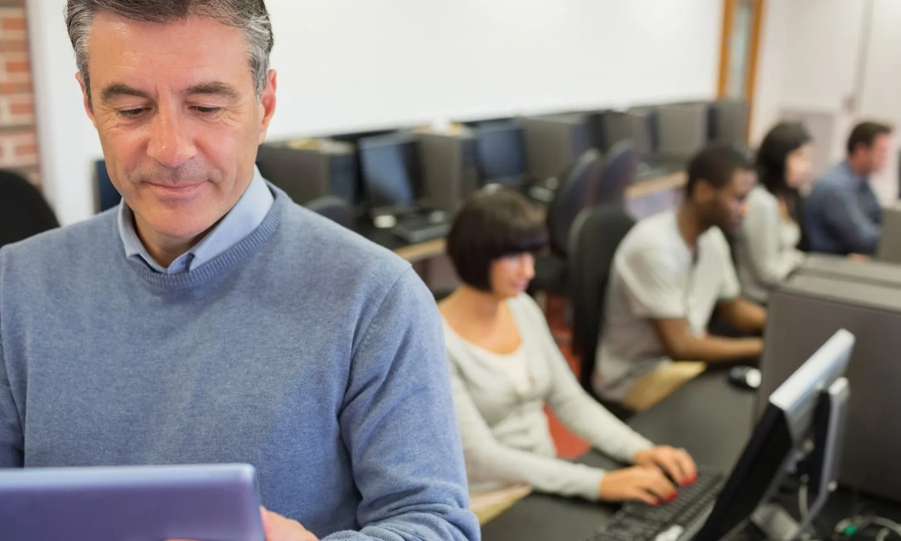 Una persona amb una tablet i al darrera podem veure diferents persones amb ordinadors.