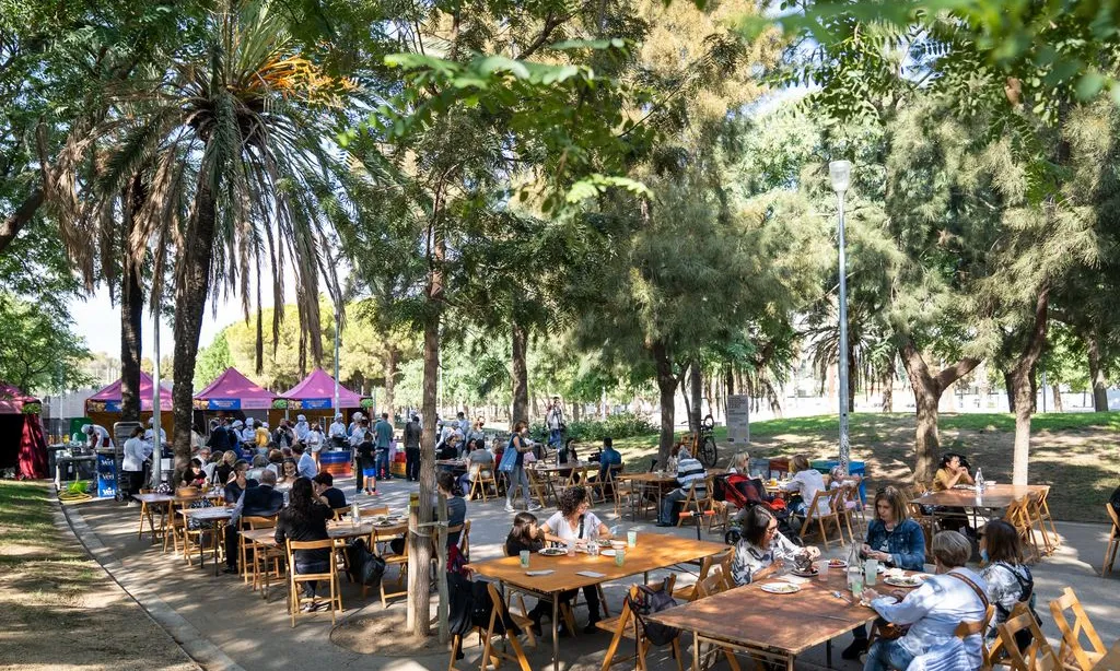Persones menjant en taules parades en una avinguda peatonal, envoltats d'arbres.