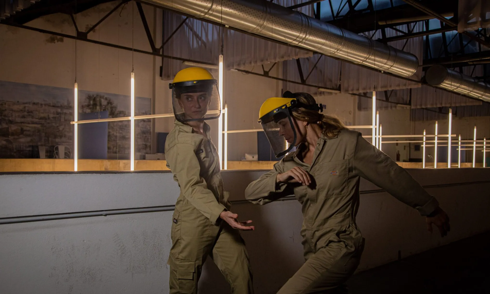 Dues persones amb roba de protecció i pantalles facials participen en una activitat dins d’un espai industrial il·luminat amb estructures metàl·liques al fons.