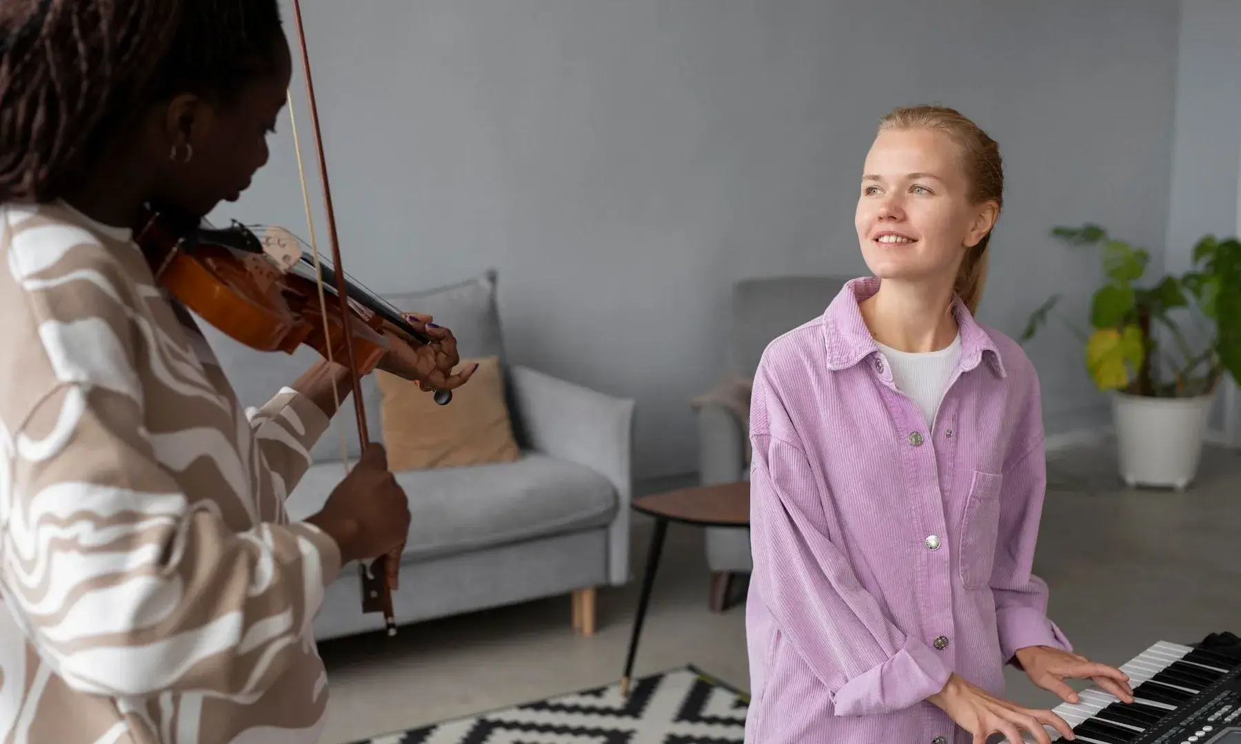 Dues persones toquen instruments musicals en un espai interior: una toca el violí mentre una altra toca un teclat, en un ambient relaxat i lluminós.