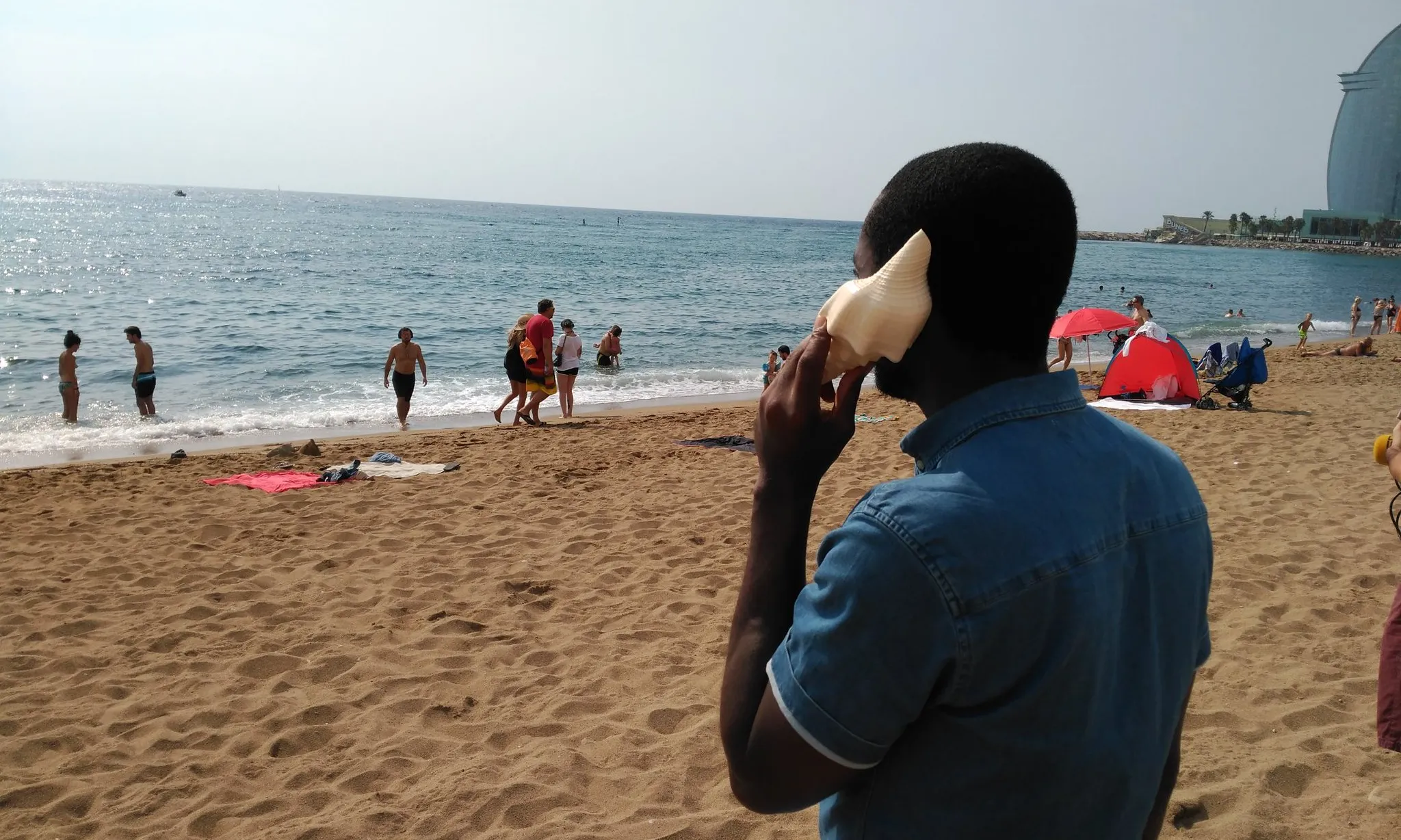 Una persona migrada s'apropa un cargol de mar a l'orella a la platja de Sant Sebastià.