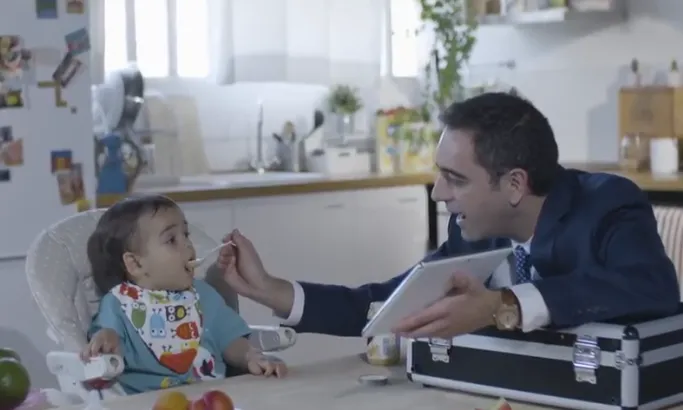 Fotograma del vídeo 'El meu primer verí'. Un home dona a menjar un potet a un nadó, mentre l'entreté amb una tauleta