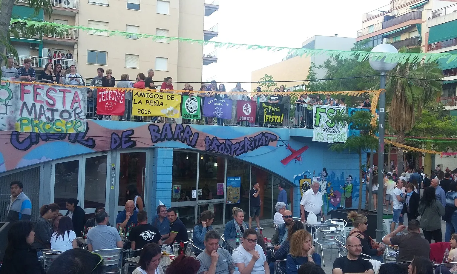 Pancartes de les penyes del barri penjades i gent asseguda a la terrassa del Casal de Barri