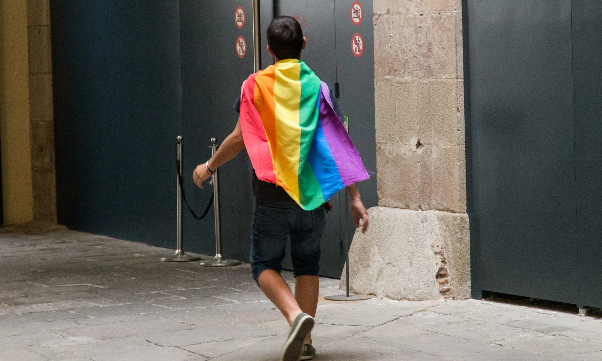 Noi jove caminant d'esquena a la càmera amb la bandera LGTBI+ penjada al coll