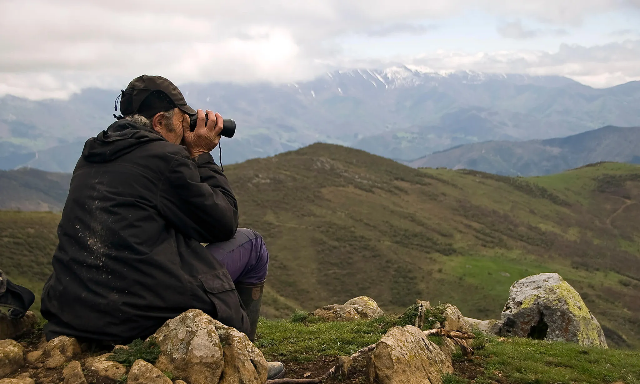 Home gran al cim d'una muntanya assegut mirant amb prismàtics el paisatge