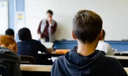 Primer pla d'un nen en una aula assegut. De fons, la docent i la pissarra. Font: Taylor Wilcox