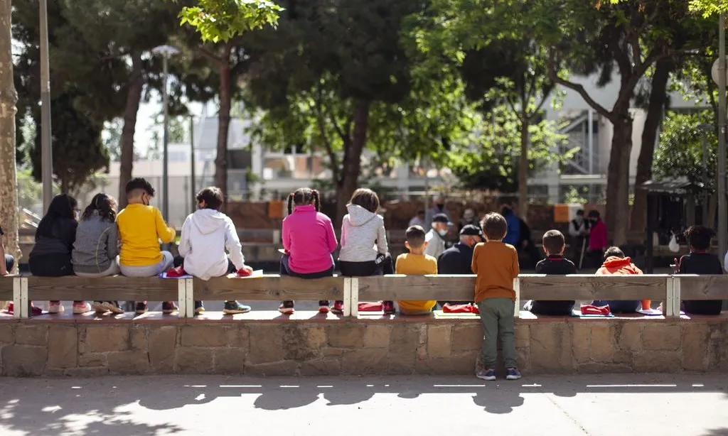 Nens de l'Escola Mercè Rodoreda asseguts en uns esglaons de la plaça de Nou Barris amb els apunts a les mans assistint a una classe.