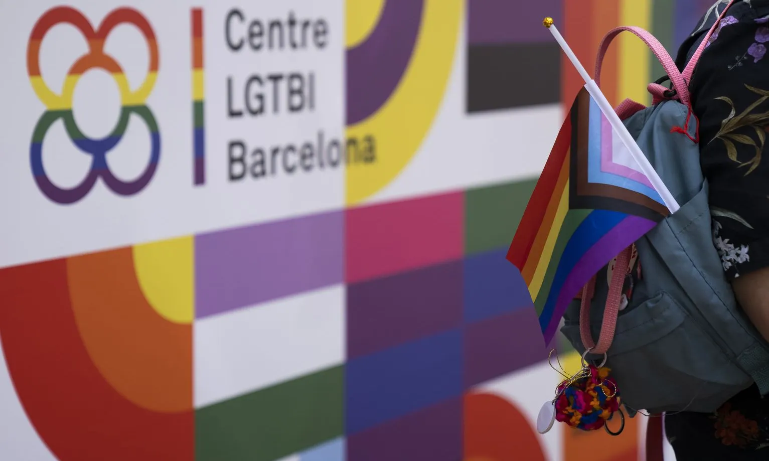 Cartell amb el logotip del Centre LGTBI de Barcelona amb una persona portant una bandera de l'arc de Sant Martí i trans enganxada a la motxilla.