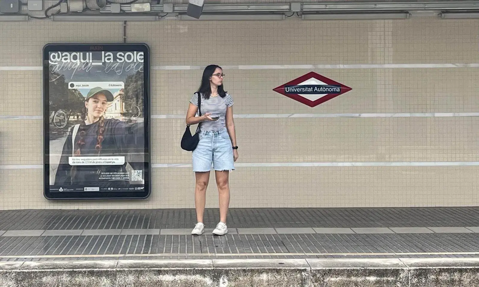 Una noia esperant el metro i, a darrere, un cartell oficial de la campanya 'Aquí la Sole'.