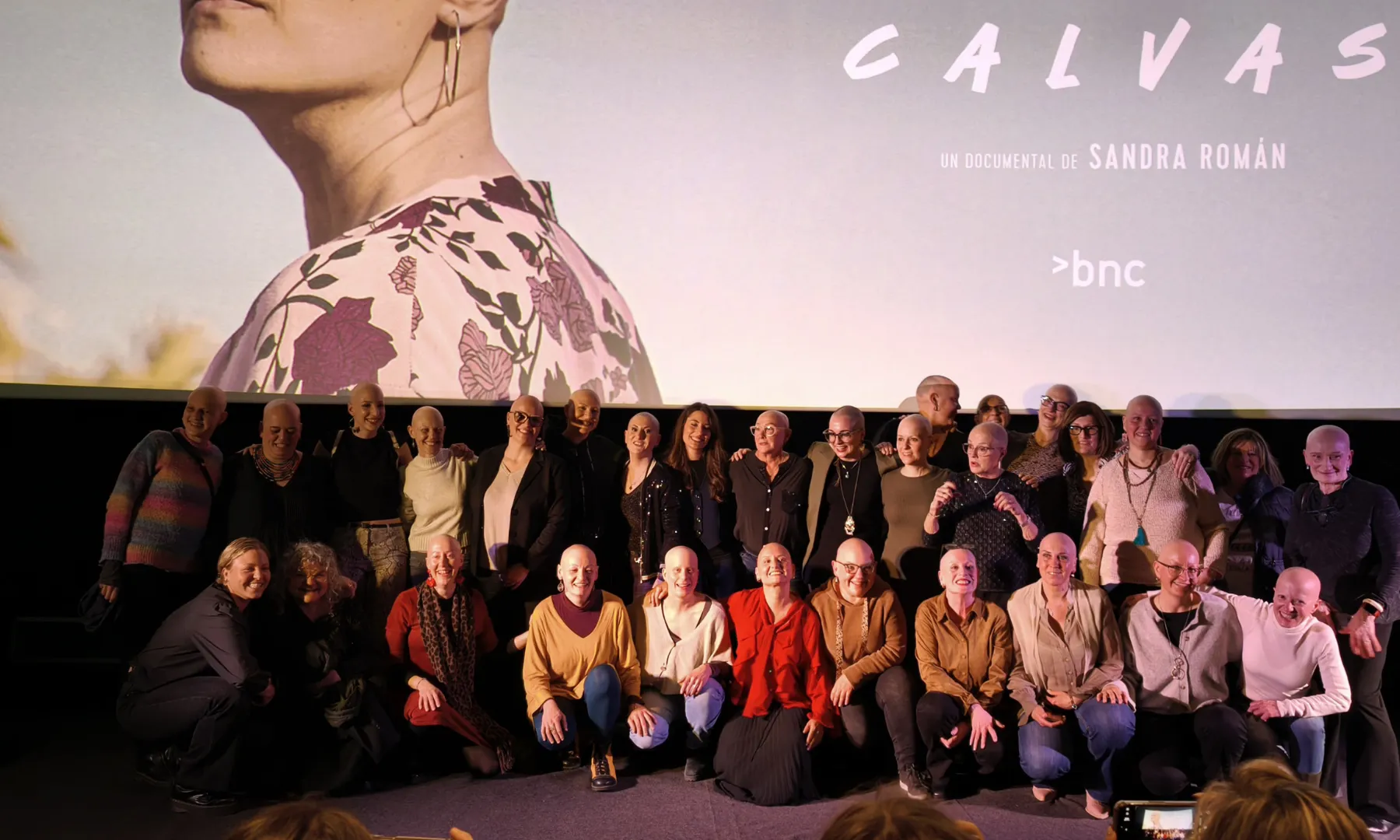 Grup de dones amb alopècia posant juntes davant d’una pantalla de cinema durant la presentació d’un documental.