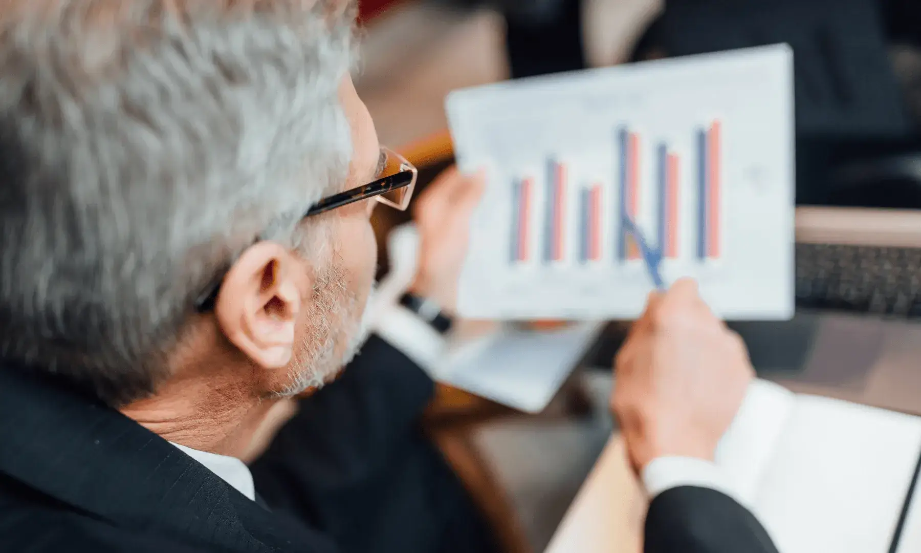 Un home amb cabells grisos i ulleres, vestit amb americana, sosté un full amb un gràfic de barres de colors i l’assenyala amb un bolígraf mentre està assegut davant d’un ordinador portàtil en un entorn de reunió.