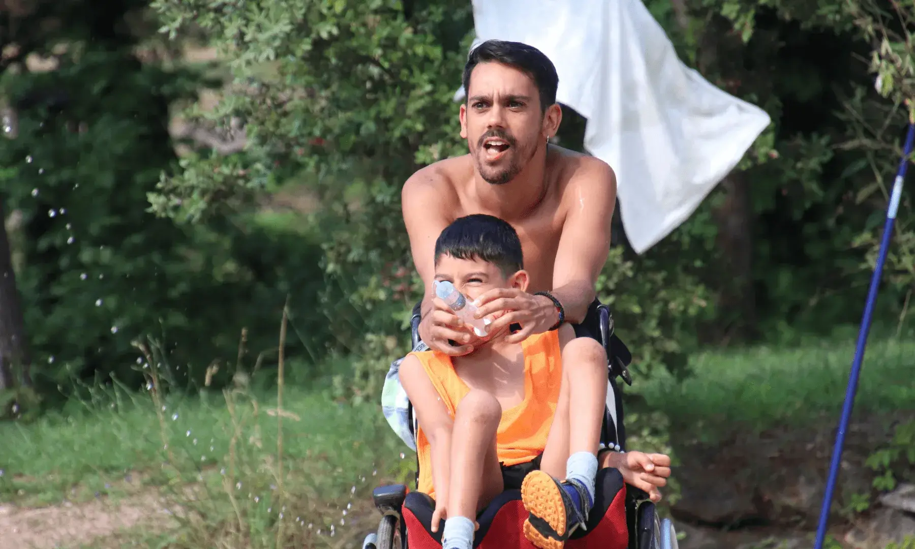 Monitor acompanya un infant amb mobilitat reduïda en cadira de rodes mentre juguen amb aigua en un entorn natural.