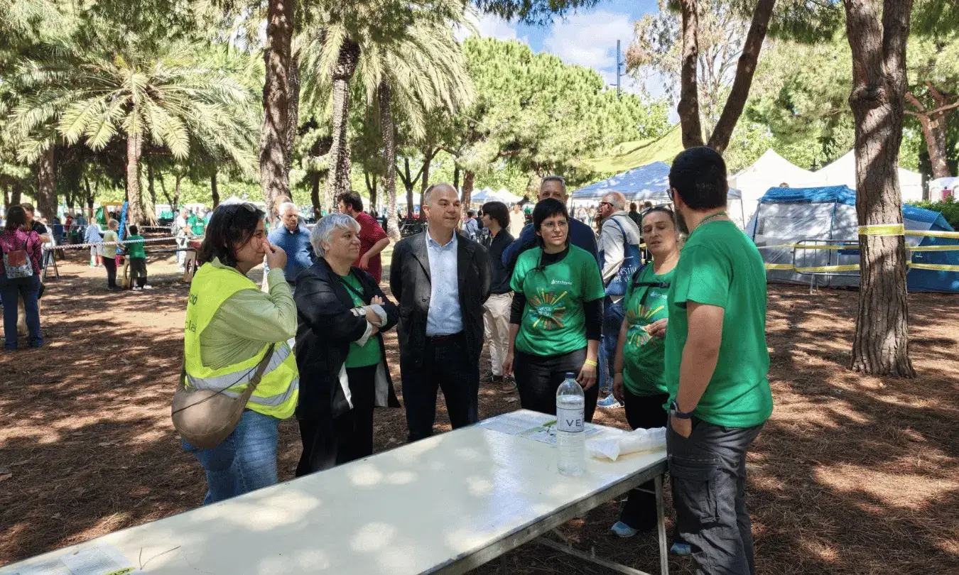 Un grup de persones, algunes amb samarretes verdes de Fundesplai, conversen al voltant d'una taula en un espai exterior envoltat d'arbres durant la Festa Esplai al Parc Nou del Prat de Llobregat.