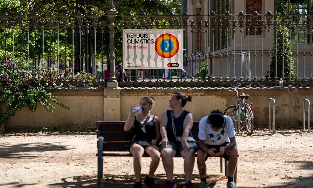 Tres persones assegudes en un banc a davant d'un parc amb un distintiu de refugi climàtic.