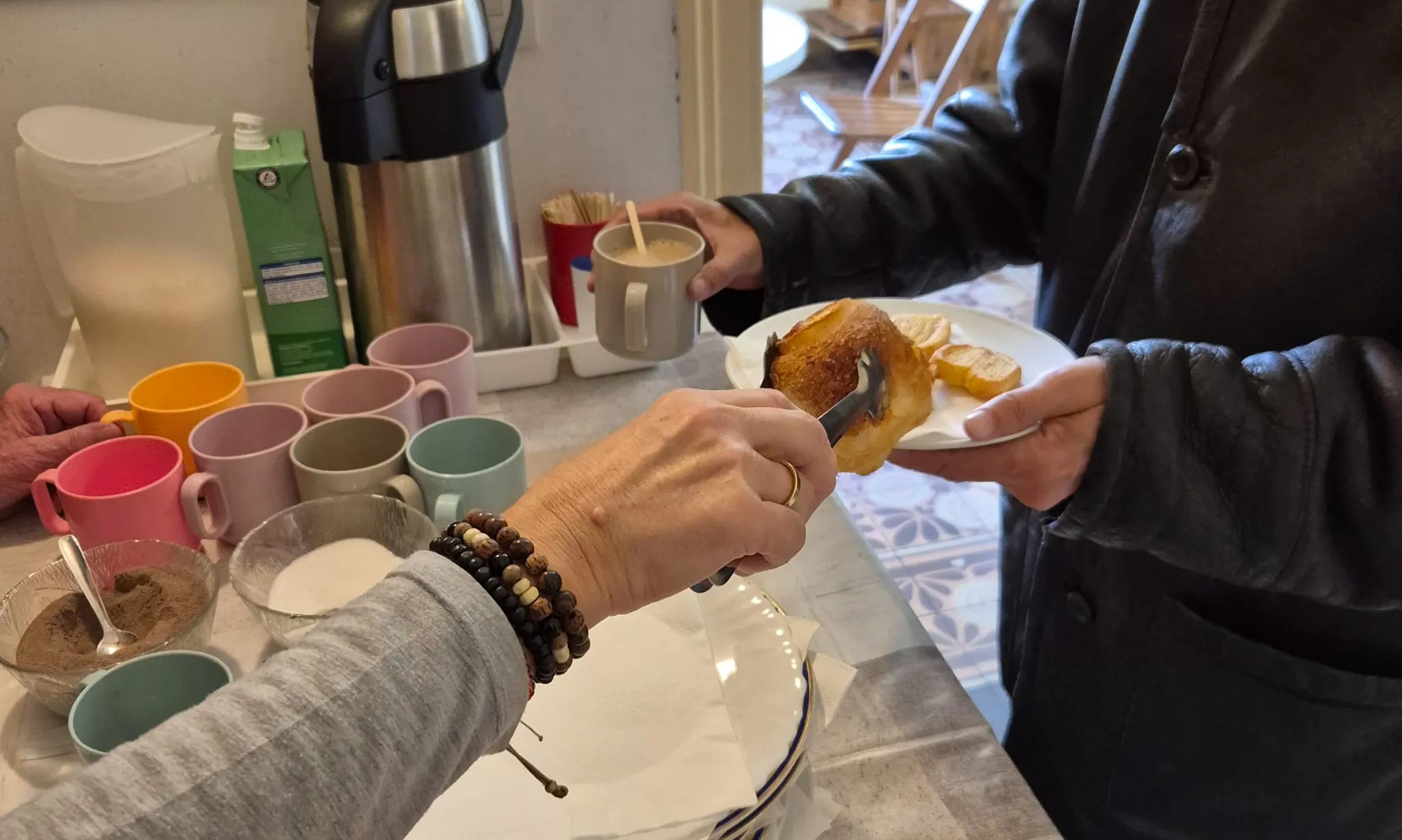 Una mà li ofereix a una altra una pasta sobre un plat, al damunt d'una taula amb un termo de café.