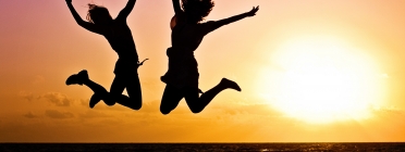 Dues noies salten d'alegria a la platja amb una posta de sol.