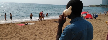 Una persona migrada s'apropa un cargol de mar a l'orella a la platja de Sant Sebastià.