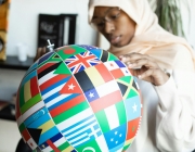 Noia mirant un globus terraqüi amb banderes del món. Font: Pexels - Monstera Production