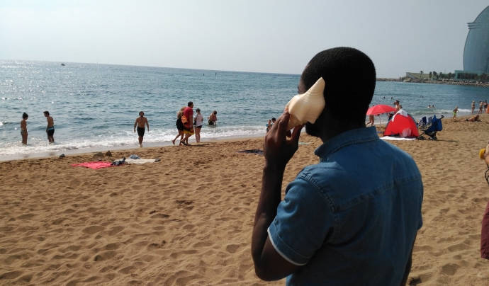 Una persona migrada s'apropa un cargol de mar a l'orella a la platja de Sant Sebastià.