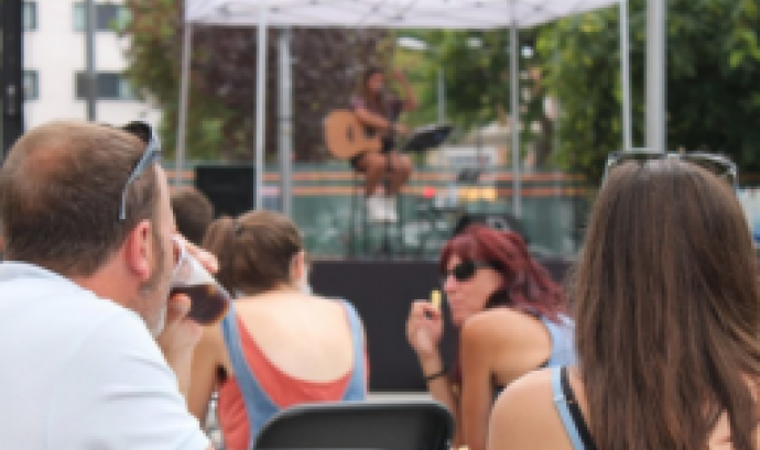 Persones assegudes d'esquena mirant al escenari, on hi ha una artista asseguda a un taburet i amb una guitarra acústica recolzada als braços
