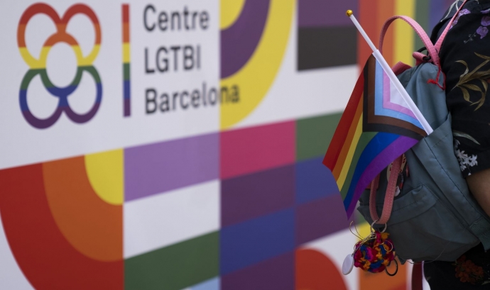 Cartell amb el logotip del Centre LGTBI de Barcelona amb una persona portant una bandera de l'arc de Sant Martí i trans enganxada a la motxilla. Font: Paula Jaume