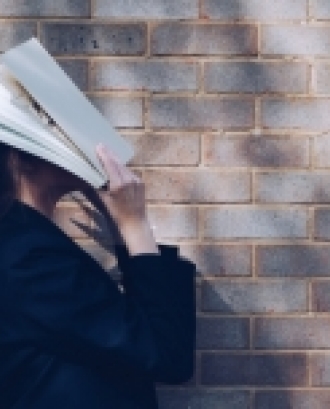 Noia tapant-se la cara amb un llibre davant d'una paret. Font: Siora